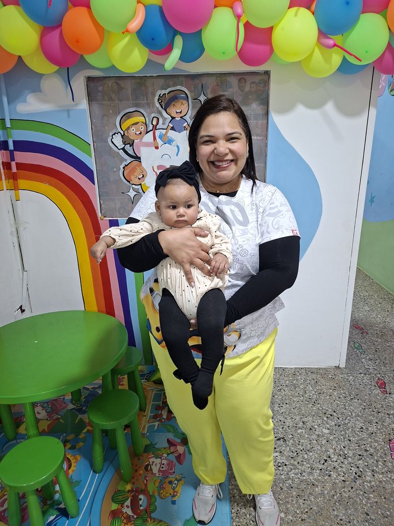 La Dra. Mine sonriendo con una bebé en brazos frente al mural del consultorio