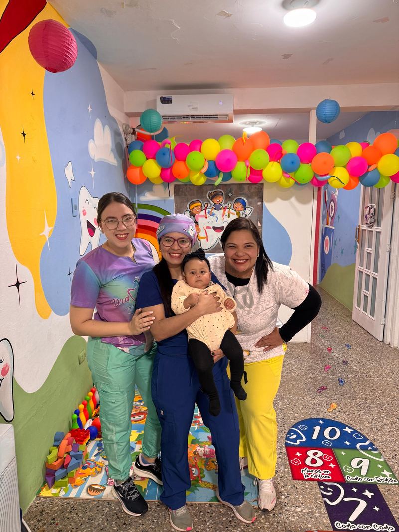 Tres integrantes del equipo del consultorio con una bebé en brazos, bajo una guirnalda de globos