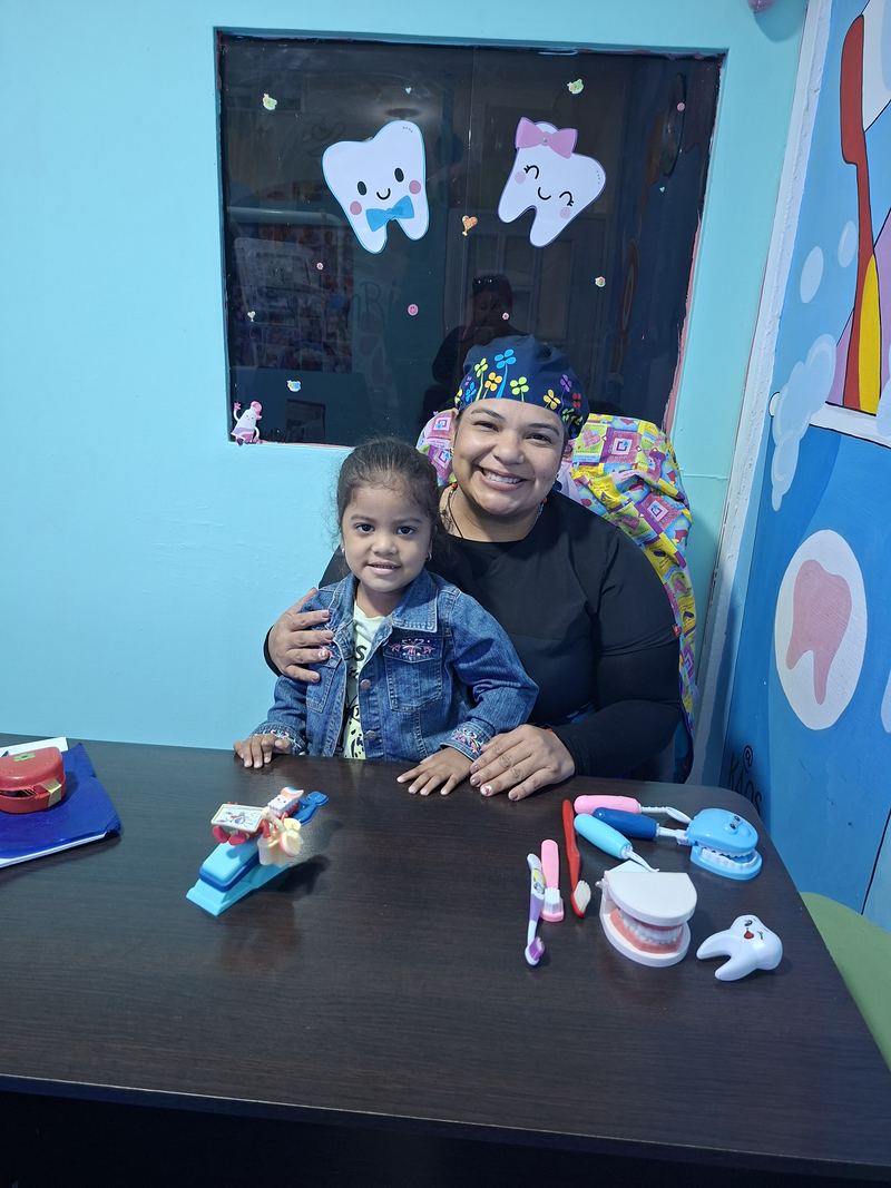 La Dra. Mine con una niña que sonríe en el escritorio, rodeadas de juguetes dentales