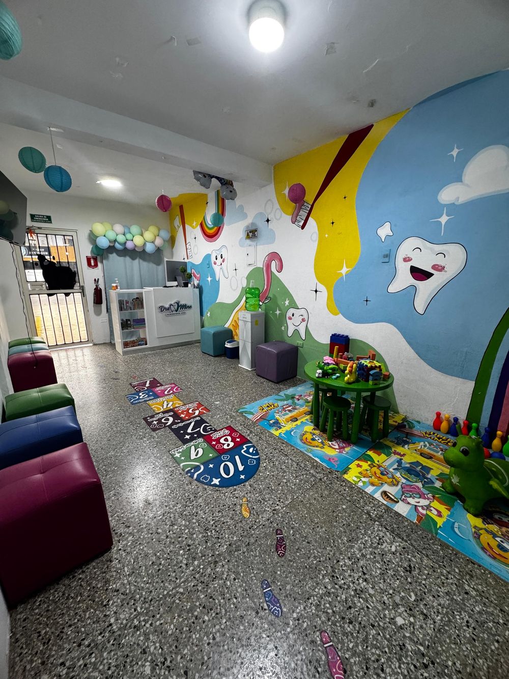 Sala de espera del consultorio con mural de dientes sonrientes, rayuela en el piso y rincón de juegos